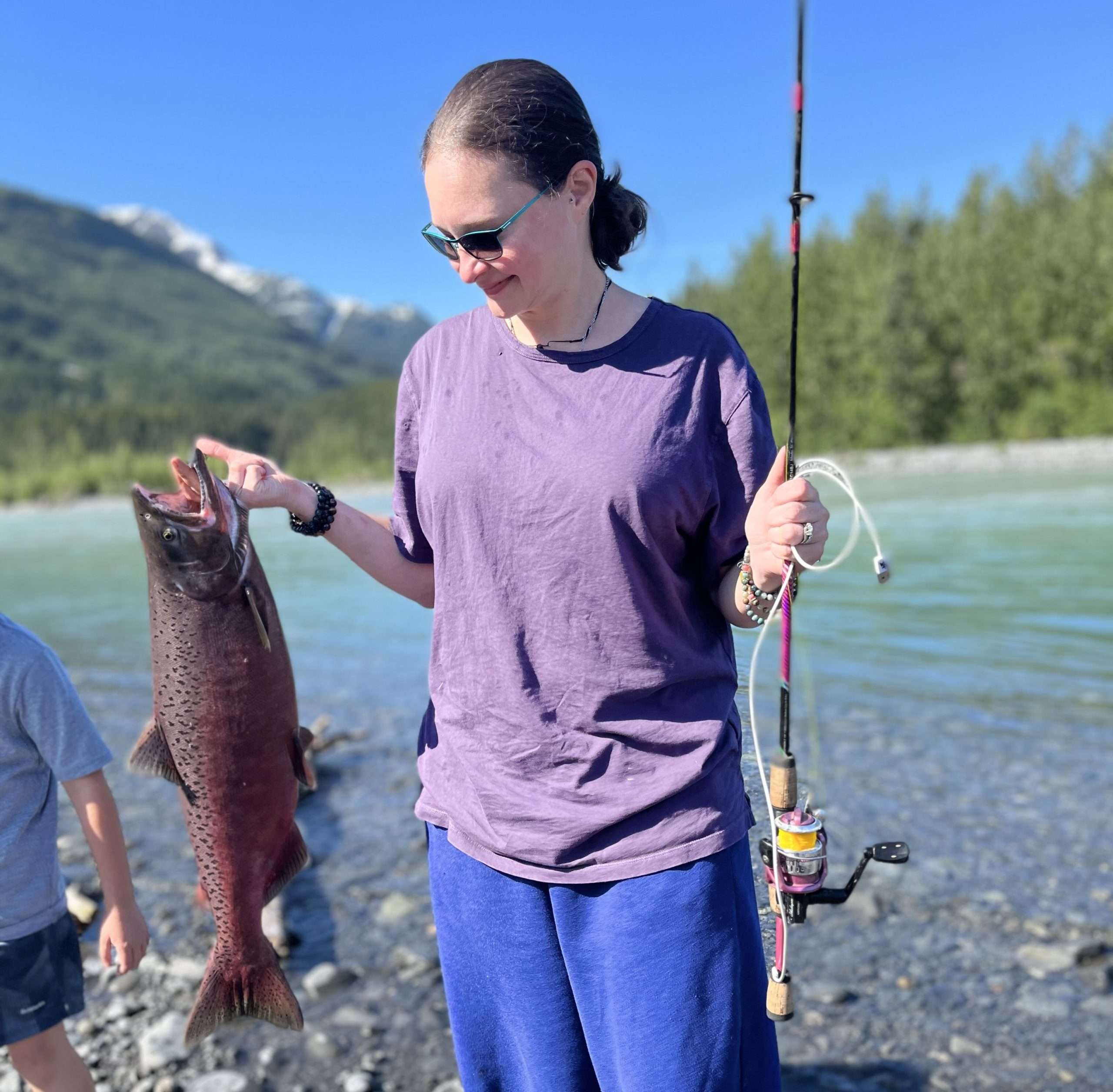 Person fishing by a scenic river, holding a large catch, sunny day.